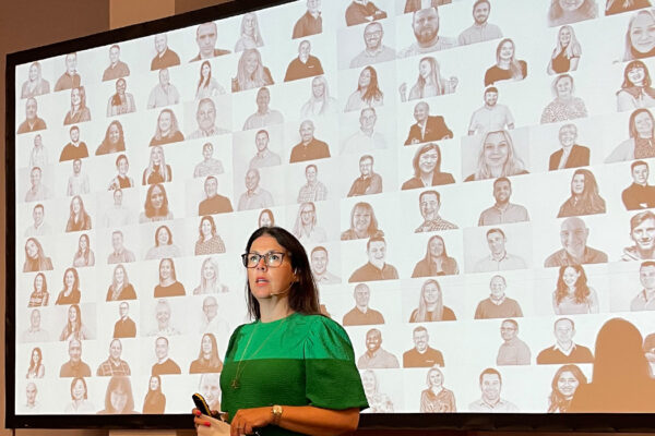 Brunette woman in green shirt with glasses, holding remote control and paper, wearing lavalier microphone. Projector screen with collage of headshot photographs behind her.