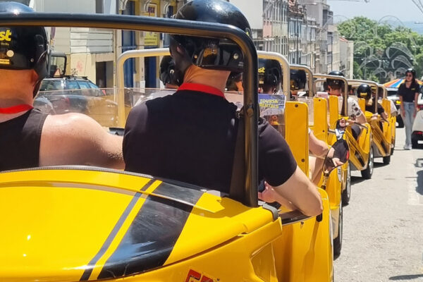 A group of six brightly-yellow coloured go karts riding single file in a town setting. There are two people to a go kart, and each person wears a helmet.