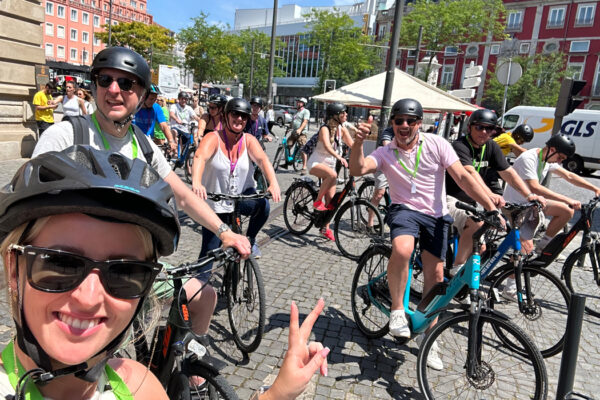 Smiling woman's selfie of eight people on electric bicycles in a town. Man on right gives 'thumbs up.' Everyone wears helmets. Background filled with other cyclists.